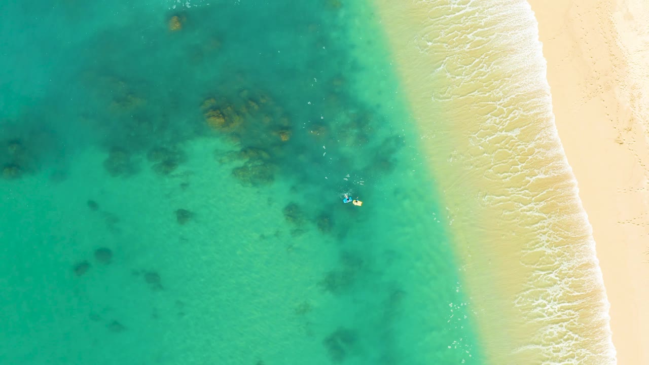 At 0:02, an overhead Shipwrecks Los Cabos view shows more open water to the left, pale sand to the right, scattered reef texture, and the tiny swimmer near the center-right shallows.