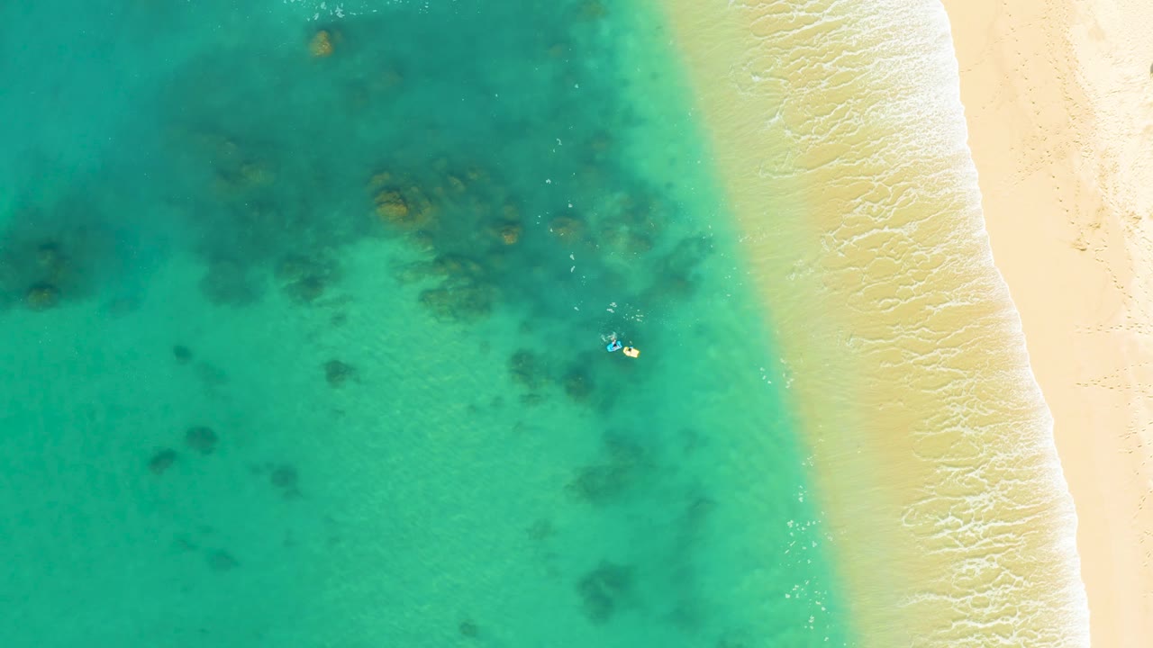 Overhead shoreline with turquoise water, reef shadows, sand, waves, and a swimmer.