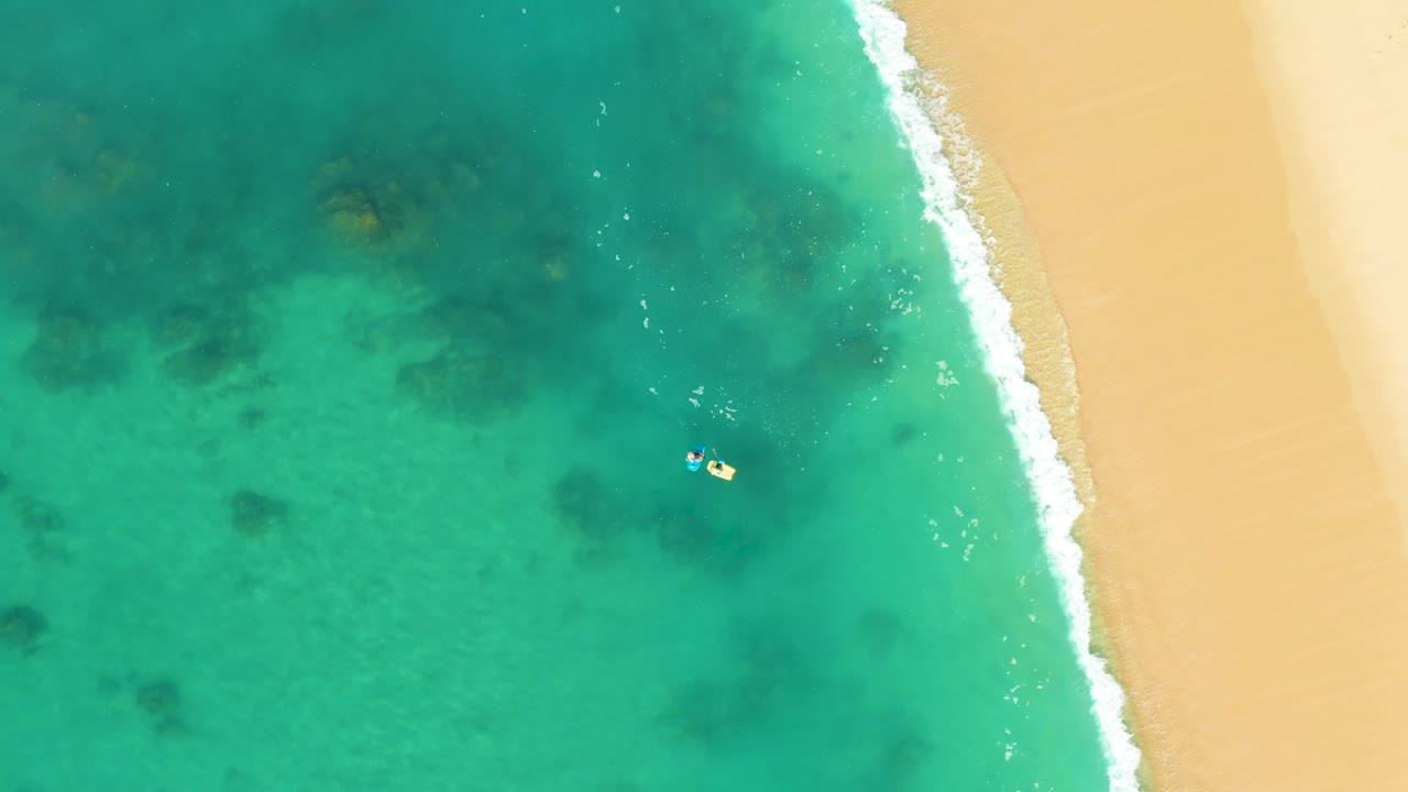 Overhead Shipwrecks Los Cabos view with the swimmers slightly right of center, scattered dark seabed formations below them, and pale sand running along the right edge.