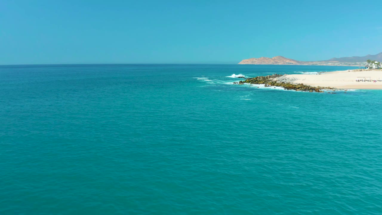 Wide aerial view at Marquis Los Cabos, Mexico, with turquoise water in the foreground, pale sand and rocks along the right edge, distant hills on the horizon, and no visible people.