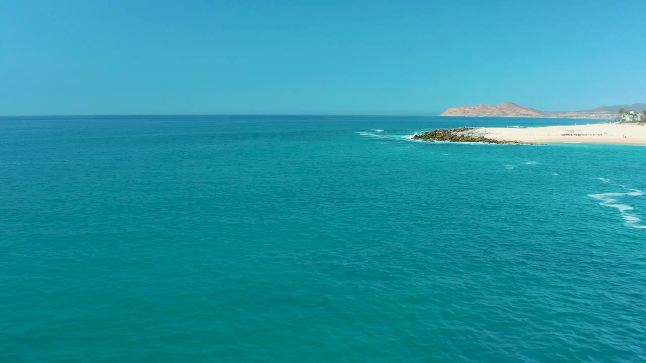 Aerial frame from Marquis Los Cabos, Mexico, showing broad turquoise water, small white foam lines, a pale sandy beach at the top right, distant hills, and no visible people.