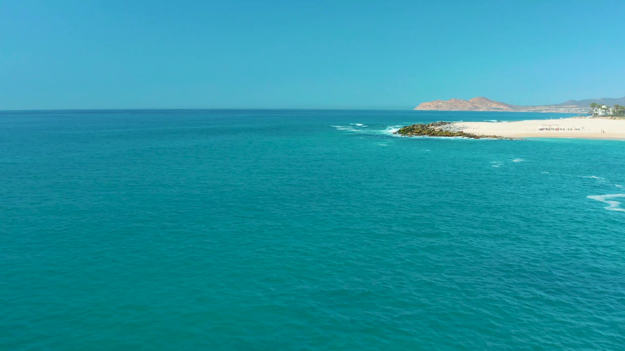 Aerial view at Marquis Los Cabos, Mexico, with rippled turquoise ocean, a curved white foam trail, a rocky edge beside the sandy beach, distant landforms, and no visible people.