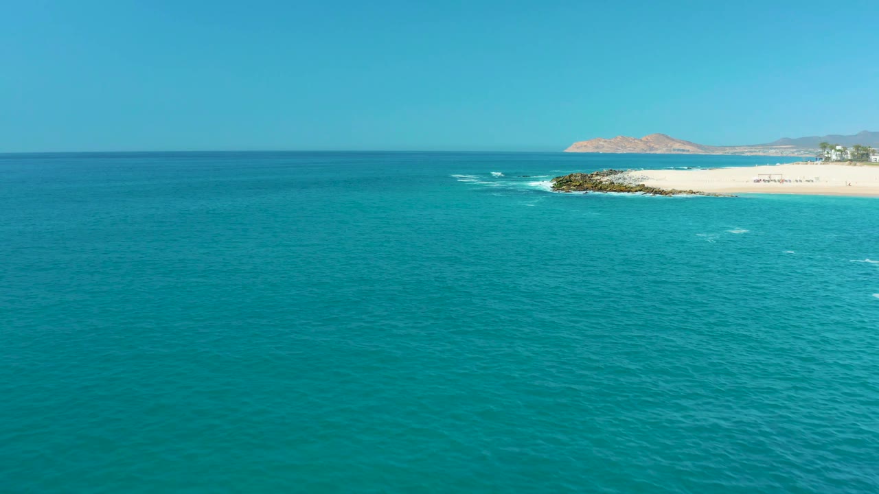 Mid-clip aerial frame at Marquis Los Cabos, Mexico, showing turquoise sea across the foreground, a bright sand strip and rocks near the upper right, faint surf breaks, and no visible people.