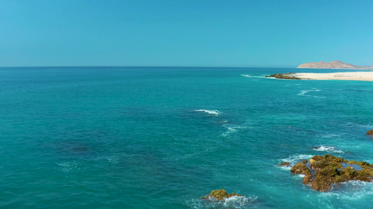 Turquoise ocean with rocks, white surf, sandy beach, and clear sky.