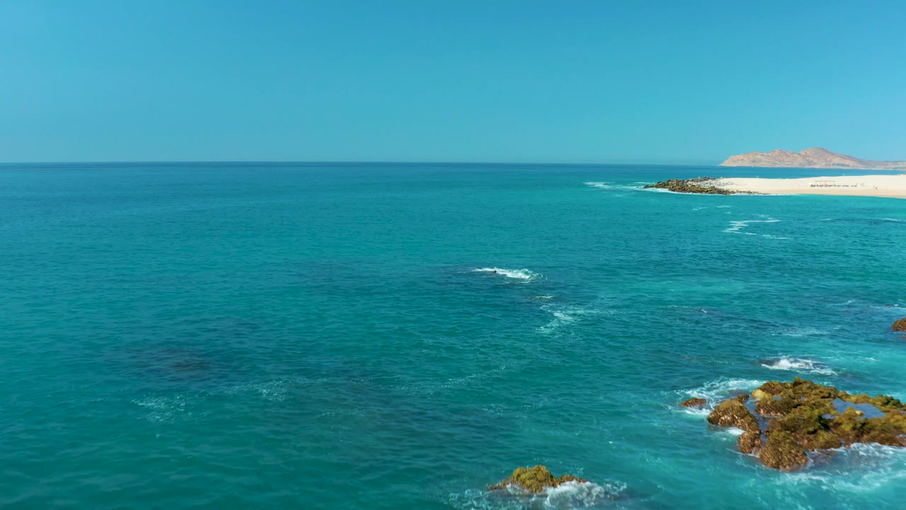 Wide elevated view over Marquis Los Cabos water, with white foam curving near midframe rocks, a pale beach in the distance, clear sky, and no visible people.