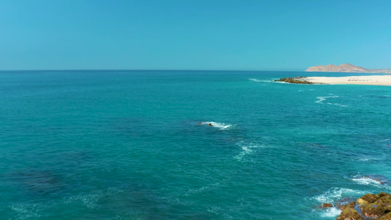 Aerial Marquis Los Cabos coastline view with the main rock cluster near the lower right edge, white surf lines across turquoise water, a sandy beach at the top right, and no visible people.