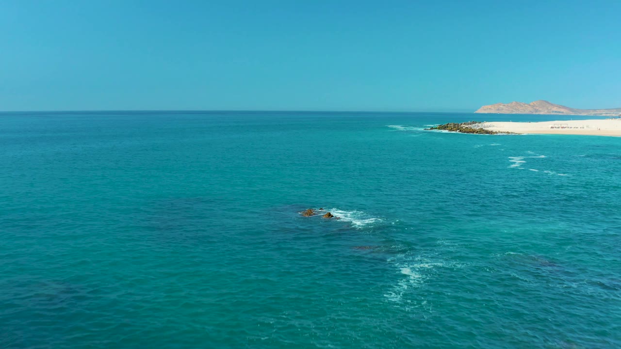 High coastal view at Marquis Los Cabos with smaller rocks near the lower center, scattered white wave marks on turquoise water, distant beach and hills, and no visible people.