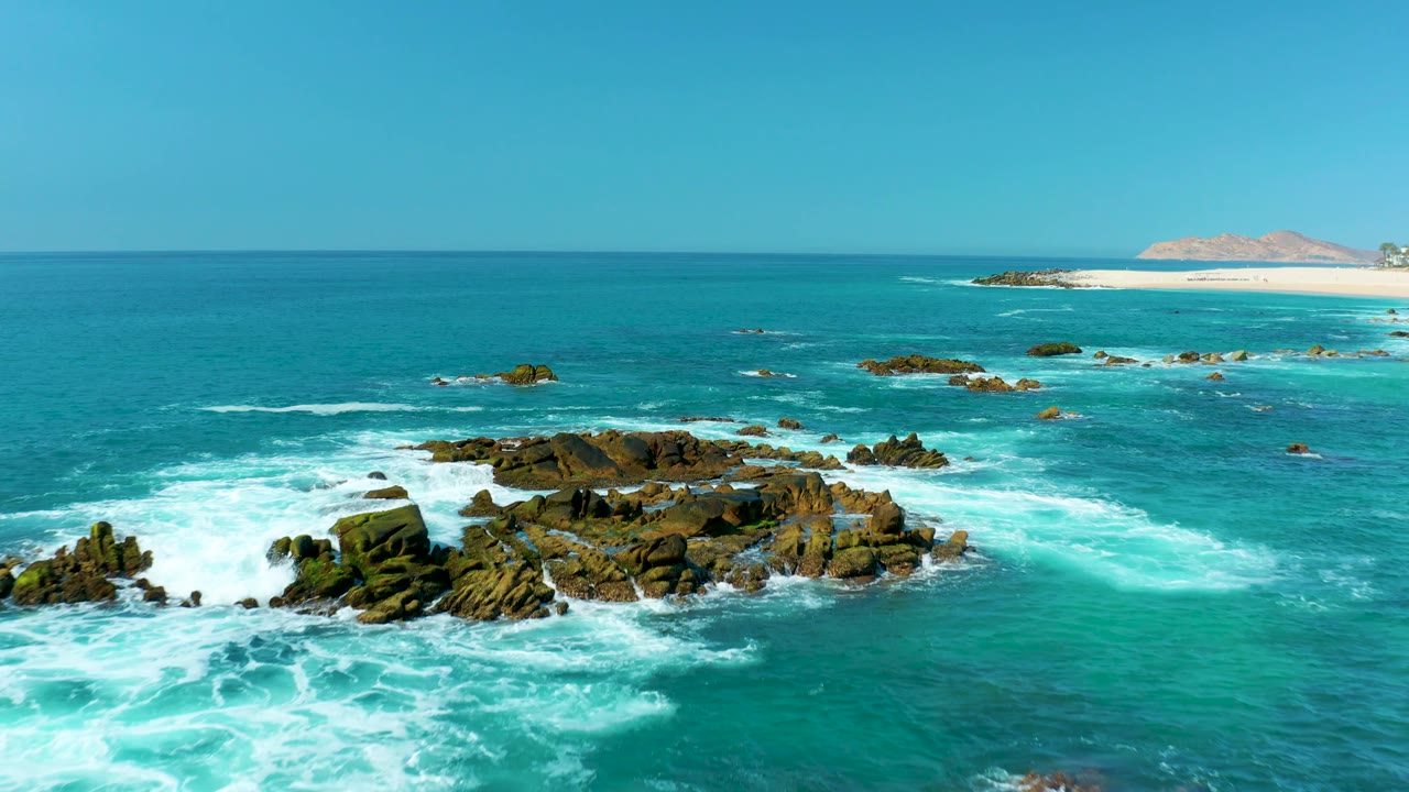 The aerial view holds on dark rocks in turquoise water as white foam spreads across the foreground, with the Marquis Los Cabos beach still visible in the far background and no visible people.