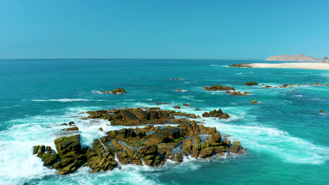 A larger rock formation fills more of the foreground as bright white surf wraps around it, with open turquoise sea, sandy beach, and distant land beyond at Marquis Los Cabos and no visible people.