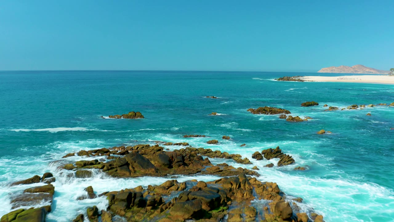 Dark rocks sit low in the foreground while white surf washes through turquoise water, with the sandy Marquis Los Cabos beach and a distant landform along the top of the frame and no visible people.
