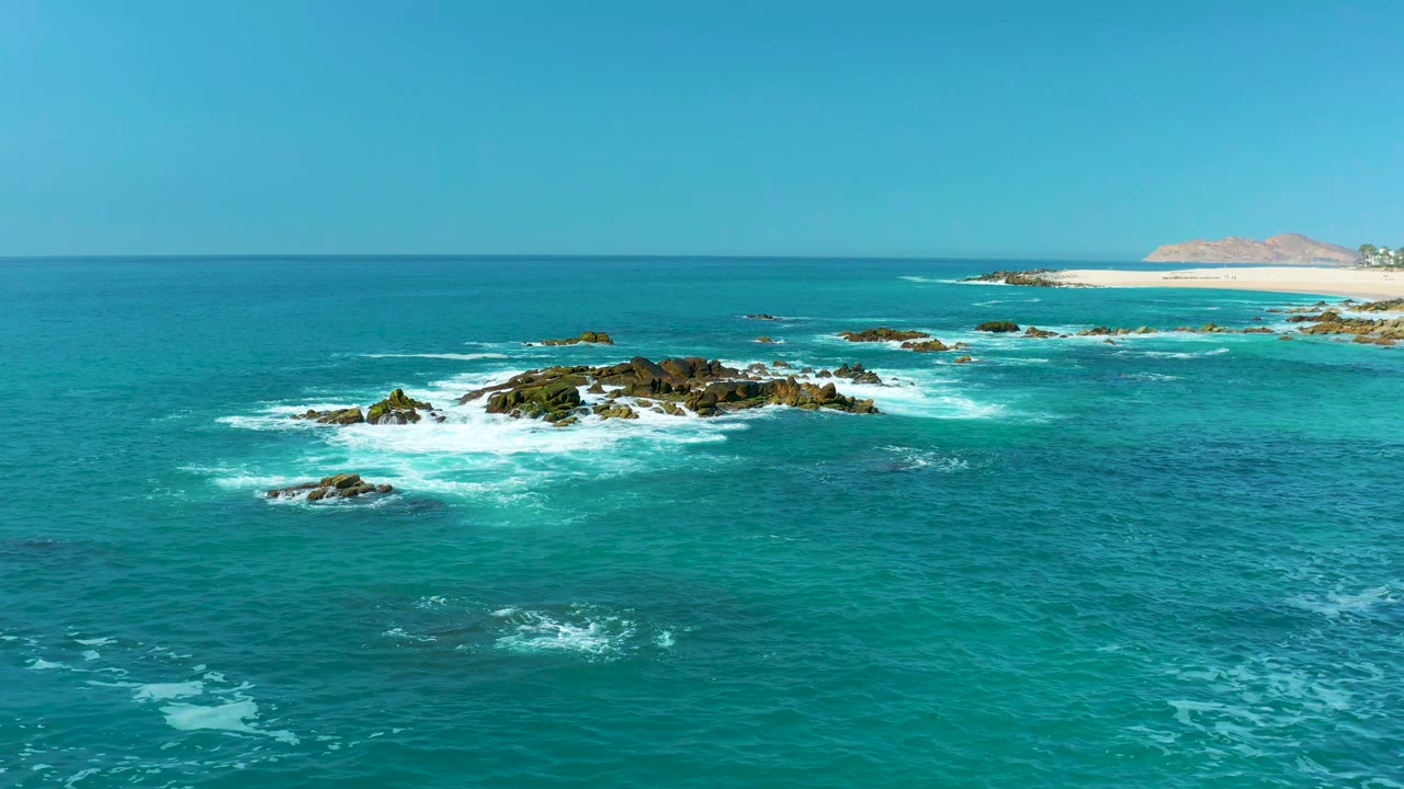 A second view of the Marquis Los Cabos shoreline shows white surf curling more tightly around the central dark rocks, with open turquoise water in front and no visible people.