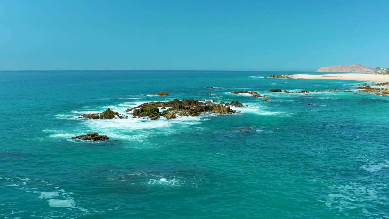 The wide elevated frame shows waves hitting dark rocks slightly left of center, teal water across the foreground, sandy beach in the rear, and no visible people.