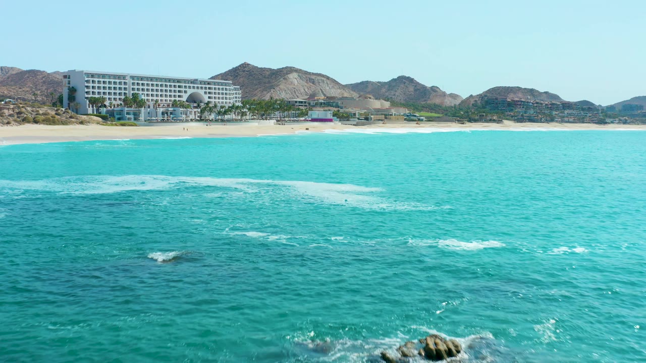 At 0:01, turquoise ocean still dominates the frame while scattered whitecaps sit lower in the water, with the Marquis Los Cabos beach, coastal building, mountains, and no visible people beyond the shore.