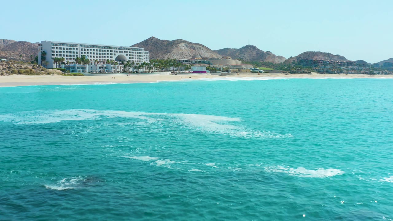 At 0:02, the water surface appears smoother on the right with small white foam patches across the foreground, while the beach, coastal building, distant mountains, and no visible people remain in view at Marquis Los Cabos, Mexico.