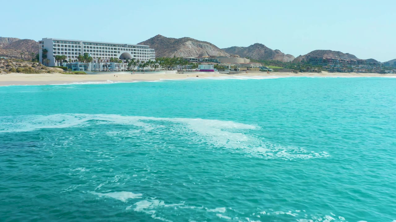 White surf line moves across turquoise water toward a sandy beach and coastal building.