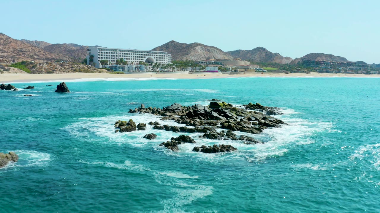 At Marquis Los Cabos, Mexico, a dark rocky outcrop sits in turquoise water with white foam, sandy beach, beachfront building, hills, blue sky, and no visible people.