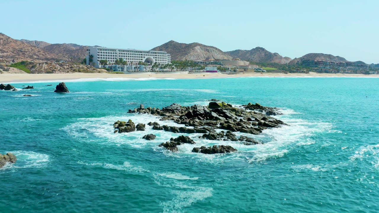 A second view at Marquis Los Cabos shows the rocky reef slightly closer in turquoise water, with white surf lines, sandy shore, large beachfront building, hills, and no visible people.