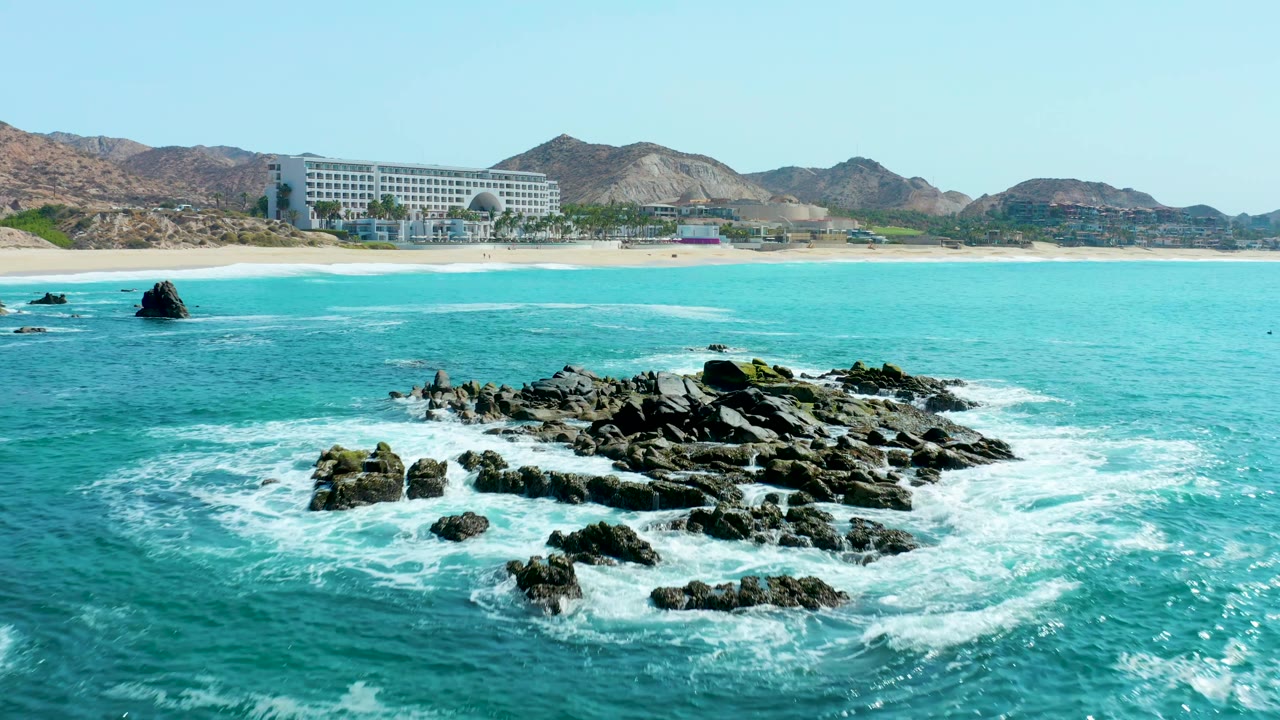 At Marquis Los Cabos, the rocky outcrop shifts toward the right side of the frame as white water curls around it, with open turquoise sea, beach, resort building, hills, and no visible people.