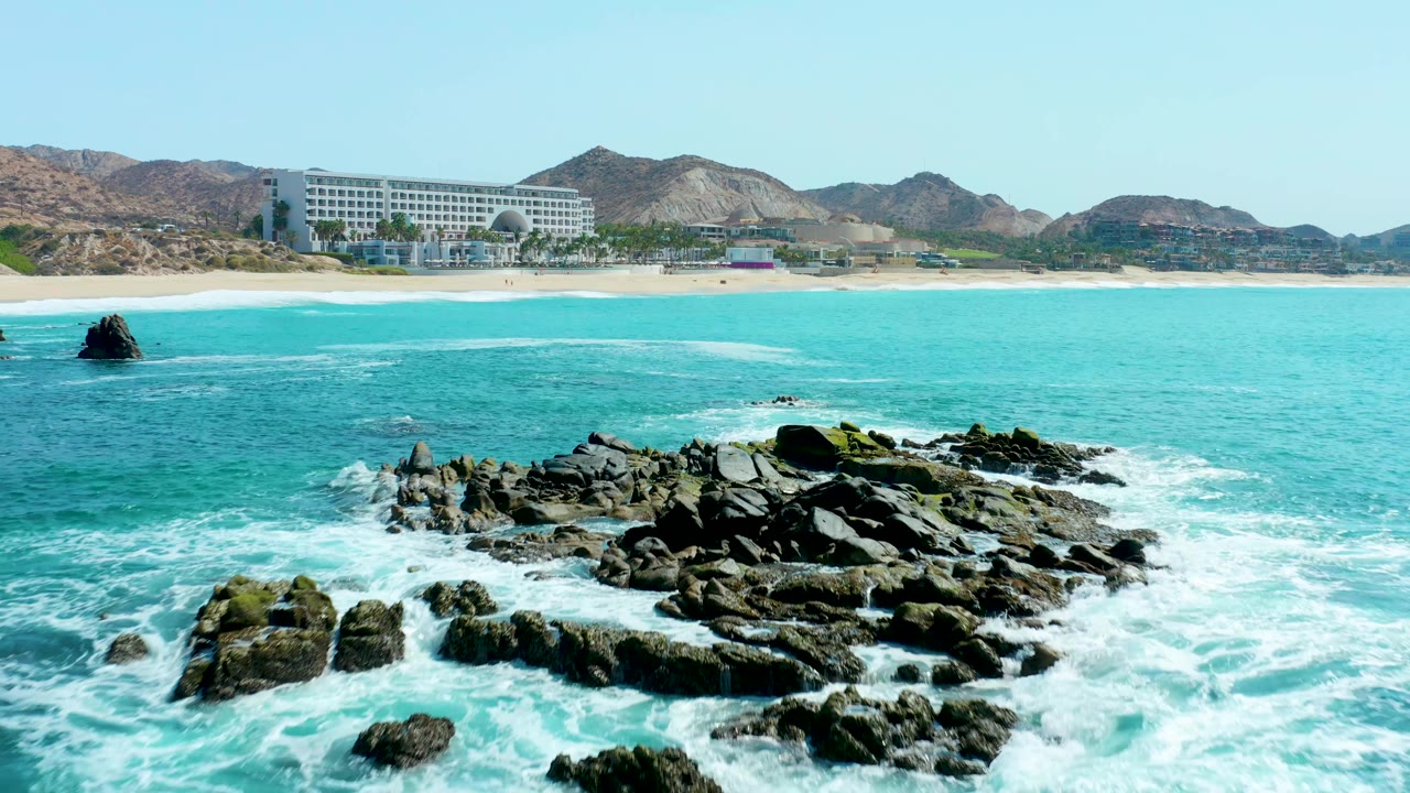 A closer Marquis Los Cabos frame shows dark foreground rocks surrounded by foamy surf, a single offshore rock at left, turquoise water, sandy beach, beachfront building, hills, and no visible people.