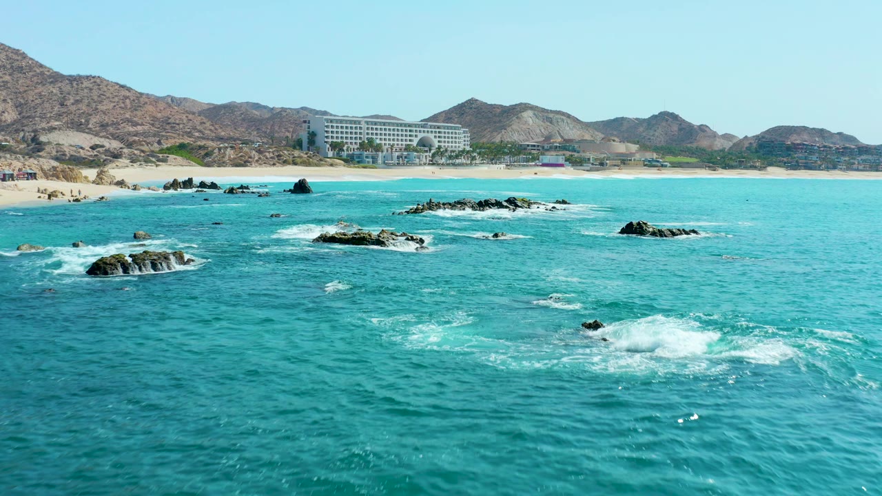 Opening view of turquoise water breaking around scattered rocks near the sandy Marquis Los Cabos beach, with a coastal building and dry mountains in the background and no visible people.