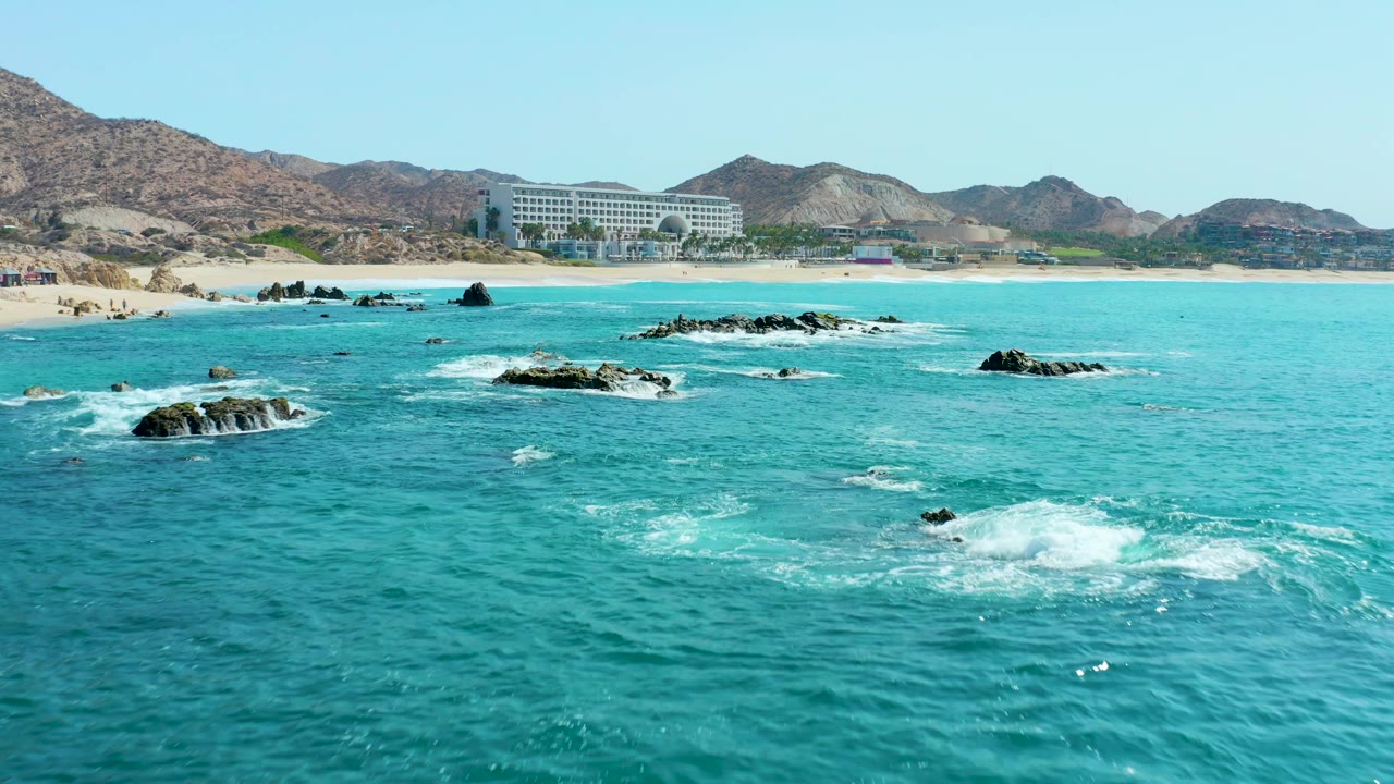 White foam spreads across turquoise water near offshore rocks and a sandy beach.