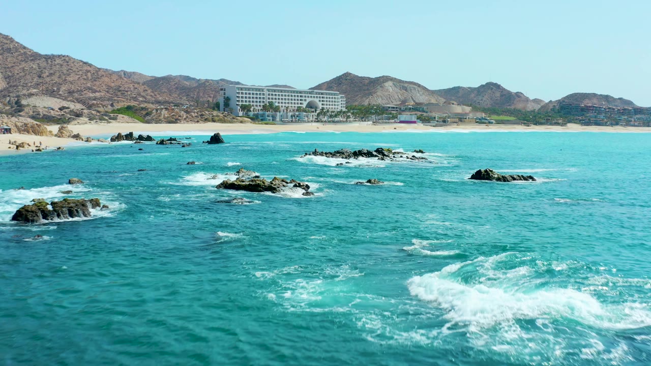 A larger patch of white surf appears at the right edge while smaller waves circle exposed rocks, with the sandy Marquis Los Cabos shoreline, resort building, and hills in the distance and no visible people.