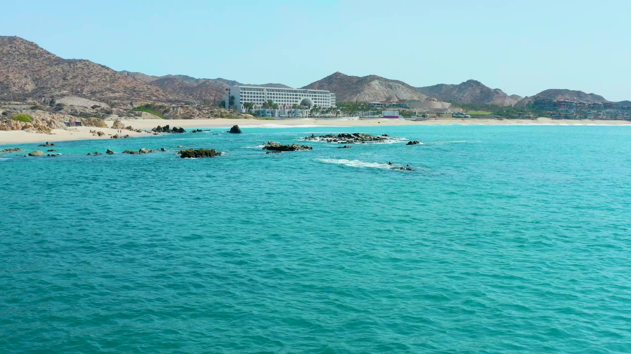 Wide aerial view at Marquis Los Cabos BCS Mexico showing turquoise water in front, scattered rock outcrops, a sandy beach, dry hills, and a large coastal building with no visible people.