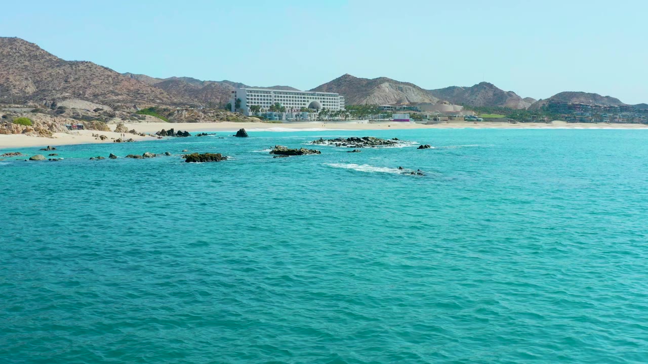 At 0:01, turquoise water covers most of the frame while a thin white wave line breaks around low rocks near the Marquis Los Cabos shoreline, with beach, hills, and a large building in back and no visible people.