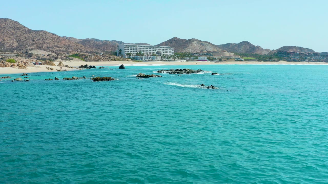 At 0:02, the rocky outcrops sit slightly farther across the midground under clear turquoise water, with the sandy Marquis Los Cabos coast, mountain ridges, and large building visible behind, and no visible people.