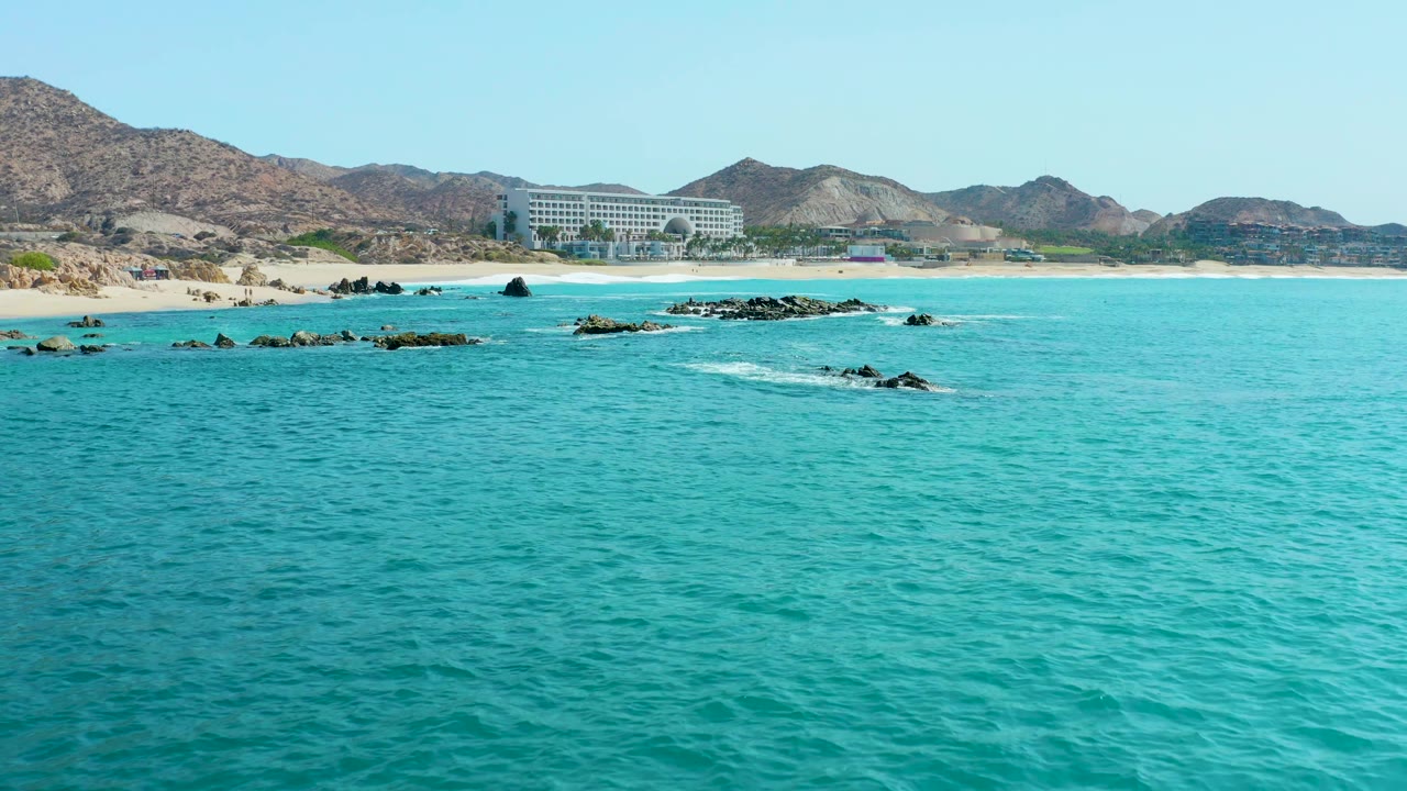 Rocky shoreline with white foam, turquoise water, beach, and distant hills.