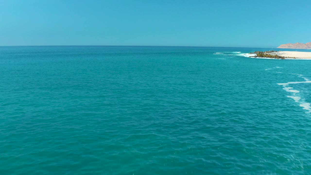 Aerial view over turquoise water at Marquis Los Cabos, Mexico, with a clear horizon, rocky shore at the upper right, white surf, and no visible people.