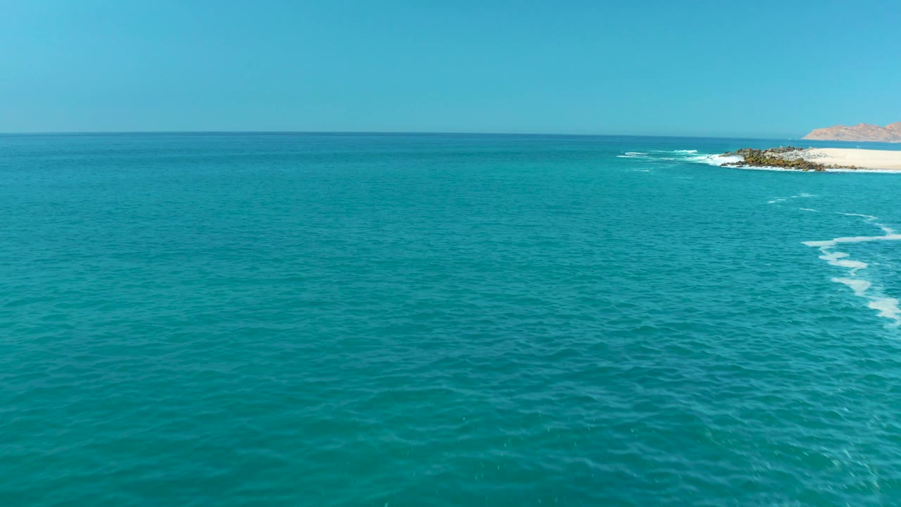 Aerial frame from Marquis Los Cabos, Mexico, showing broad turquoise sea, small white foam streaks near the right-side rocks, clear sky, and no visible people.