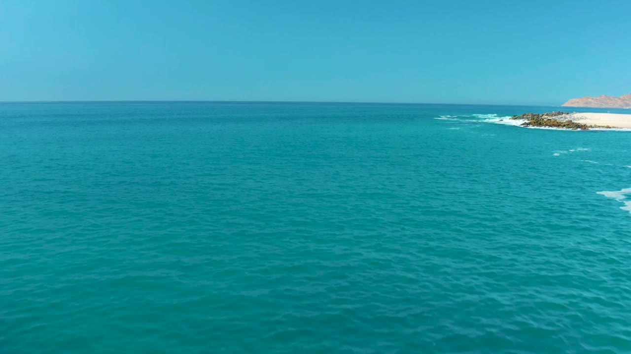 High aerial view at Marquis Los Cabos, Mexico, with open blue-green water dominating the frame, a rocky shoreline at far right, breaking white surf, and no visible people.