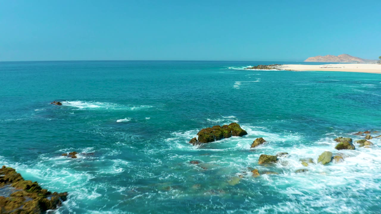 Turquoise ocean waves breaking around rocky outcrops near a distant beach.