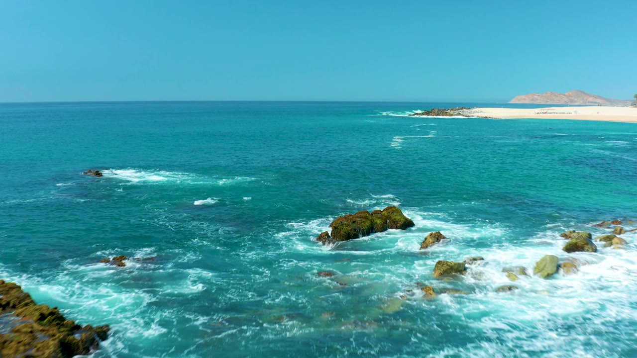 At Marquis Los Cabos, Mexico, white surf curls around clustered rocks in bright turquoise water while a sandy beach and low landmass sit along the upper horizon, with no visible people.