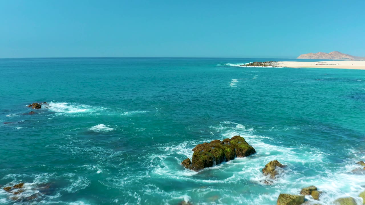 At Marquis Los Cabos, Mexico, rocky outcrops sit lower in the frame as white foam trails through turquoise water, with the distant beach visible near the horizon and no visible people.