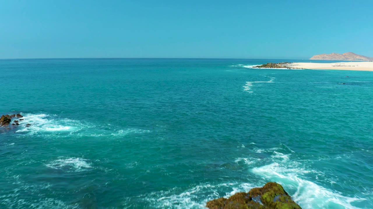 At Marquis Los Cabos, Mexico, the rocky foreground is mostly gone, leaving open turquoise water with small white surf patches, a distant sandy beach, low landmass, and no visible people.