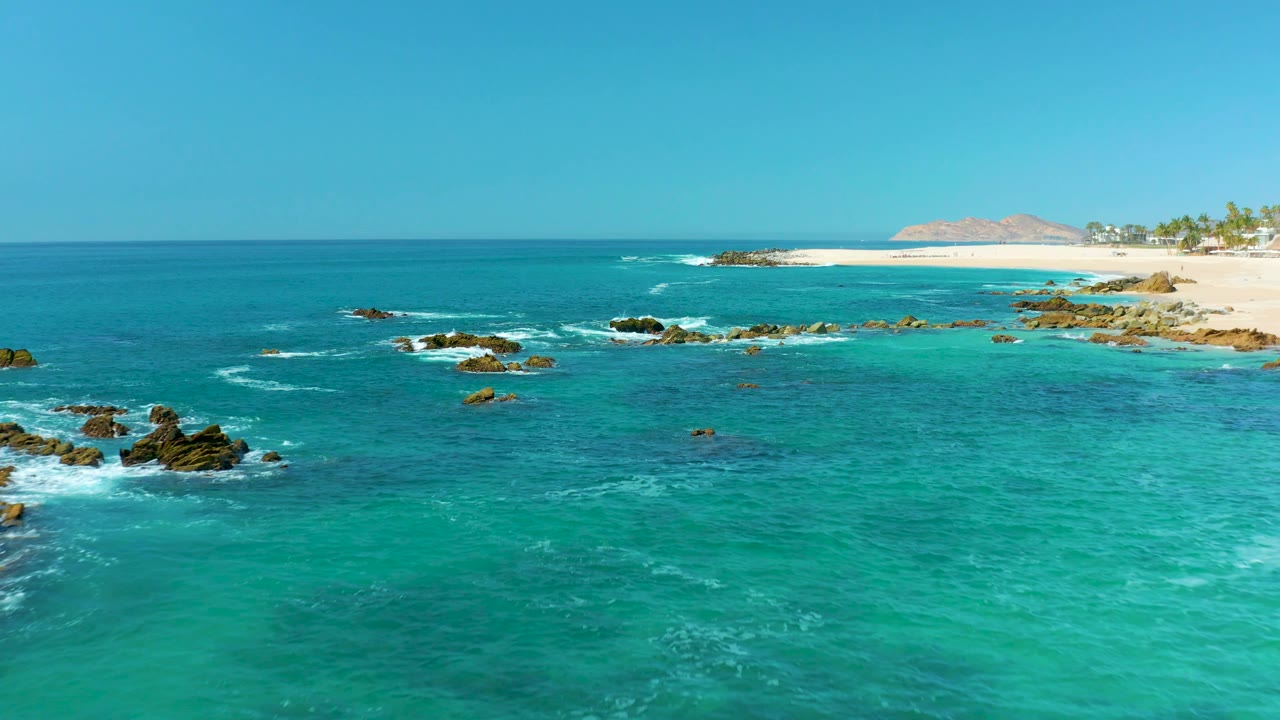 Turquoise ocean water with dark rocks, white foam, sandy beach, and distant hills.