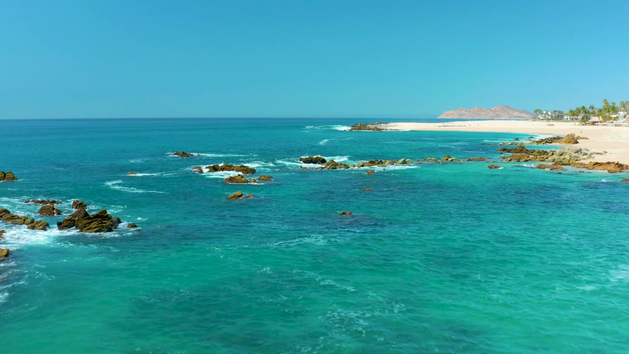 Aerial frame at Marquis Los Cabos, Mexico, showing scattered rock outcrops across clear turquoise water, thin white surf lines, a sandy beach at upper right, and no visible people.