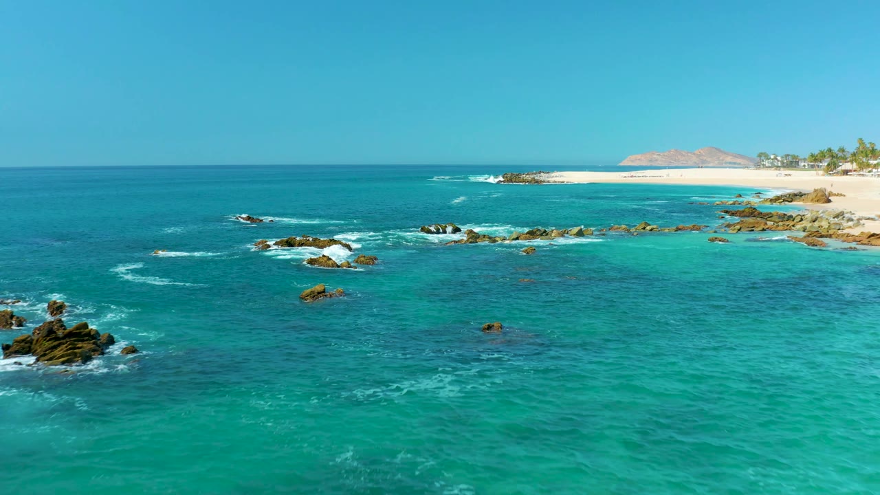 Aerial view at Marquis Los Cabos, Mexico, with the rocky surf zone shifted farther left, brighter shallow water in the foreground, sandy beach along the upper right, and no visible people.