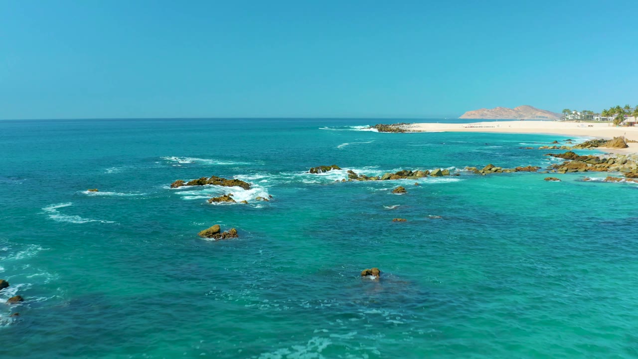 Mid-clip aerial frame at Marquis Los Cabos, Mexico, where dark rocks sit lower in the water field, white foam curls around them, the beach remains high in frame, and no visible people appear.