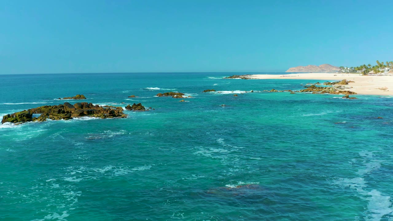 At Marquis Los Cabos, Mexico, white surf curls around a central dark rock cluster while pale sand, palms, and distant land remain visible on the upper right, no visible people.