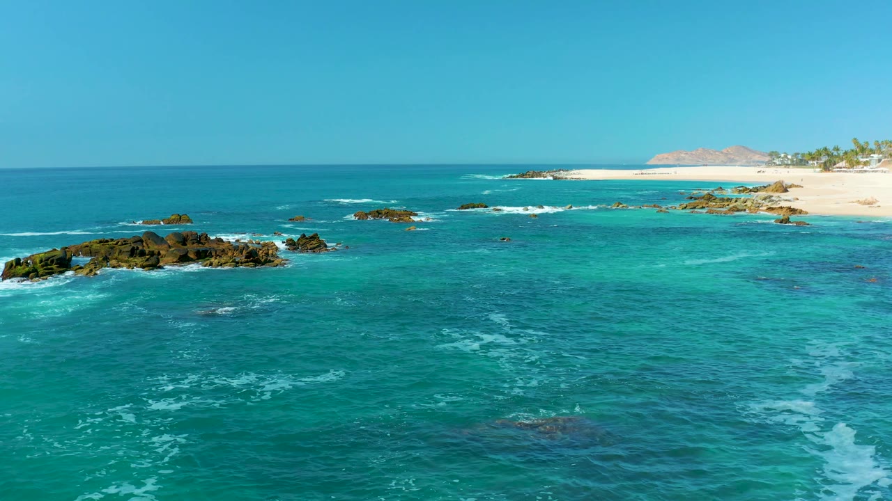 At Marquis Los Cabos, Mexico, blue-green water spreads around several low rocks with thin white wave lines and a sandy shore running across the upper right, no visible people.