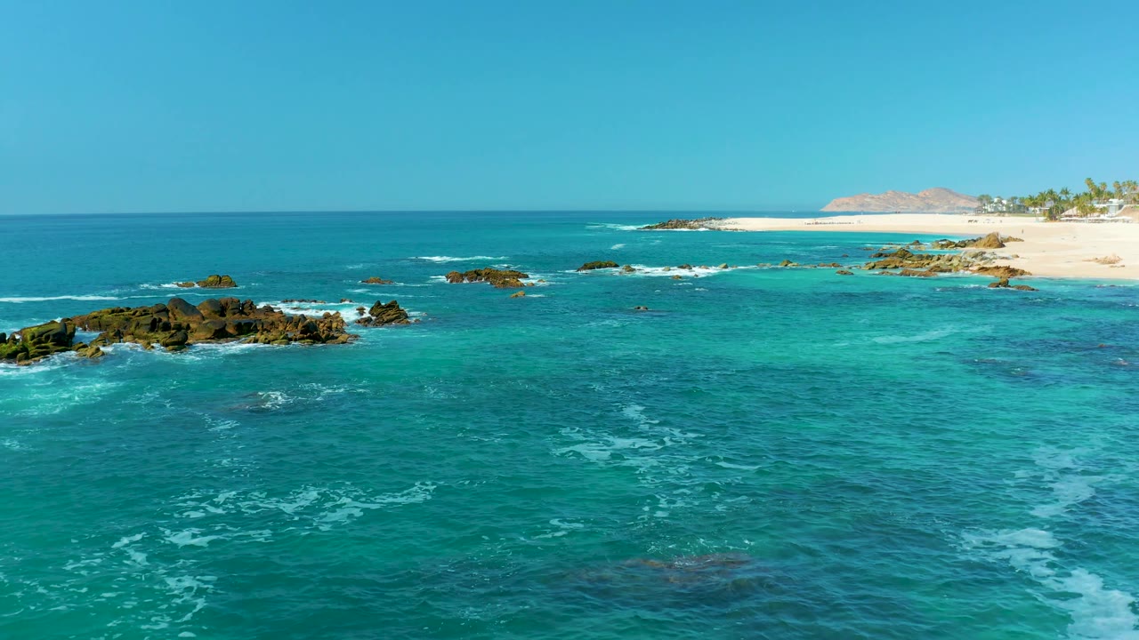 At Marquis Los Cabos, Mexico, the rocky outcrops shift lower in the frame as white surf streaks across the turquoise water below the sandy beach, no visible people.