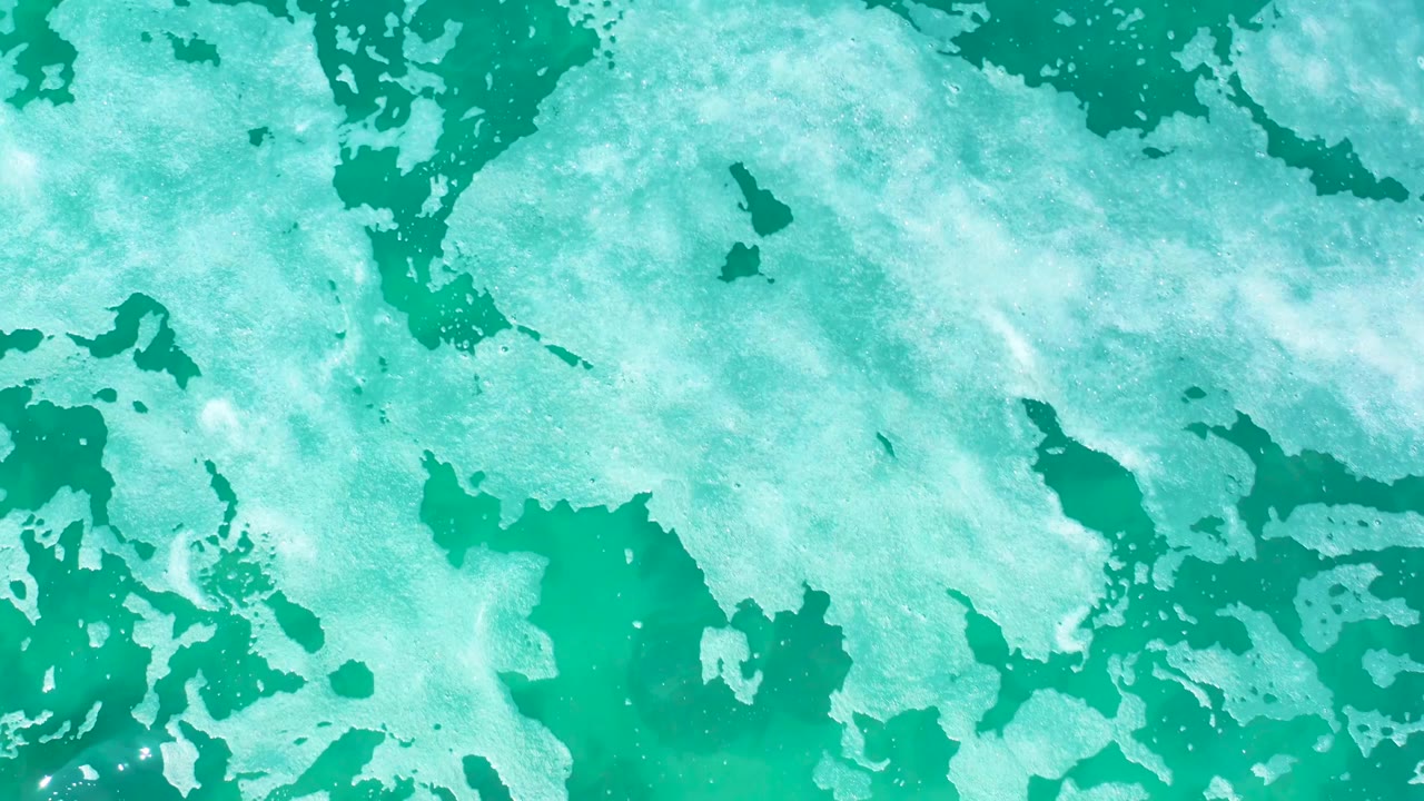 At Marquis Los Cabos in Mexico, the overhead frame shows irregular white foam shapes crossing the top and right side of the turquoise sea surface, with no visible people.