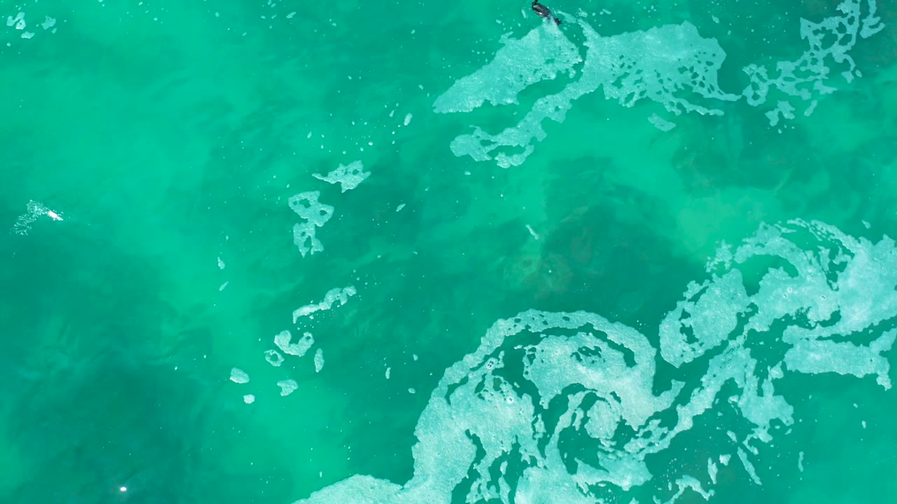 At Marquis Los Cabos, Mexico, a small swimmer appears near the upper right of a top-down turquoise ocean frame with broad white foam patches below.