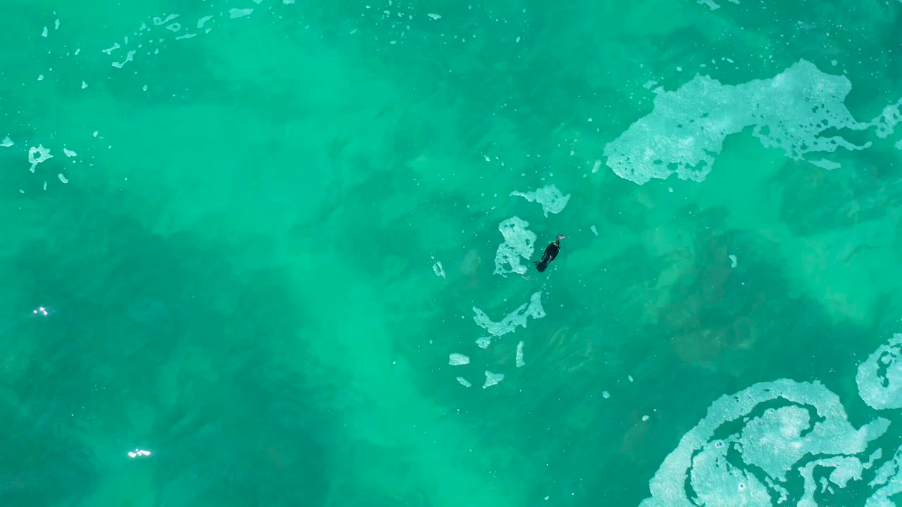 A high overhead view at Marquis Los Cabos, Mexico shows a small swimmer near pale foam patches in clear turquoise ocean water.