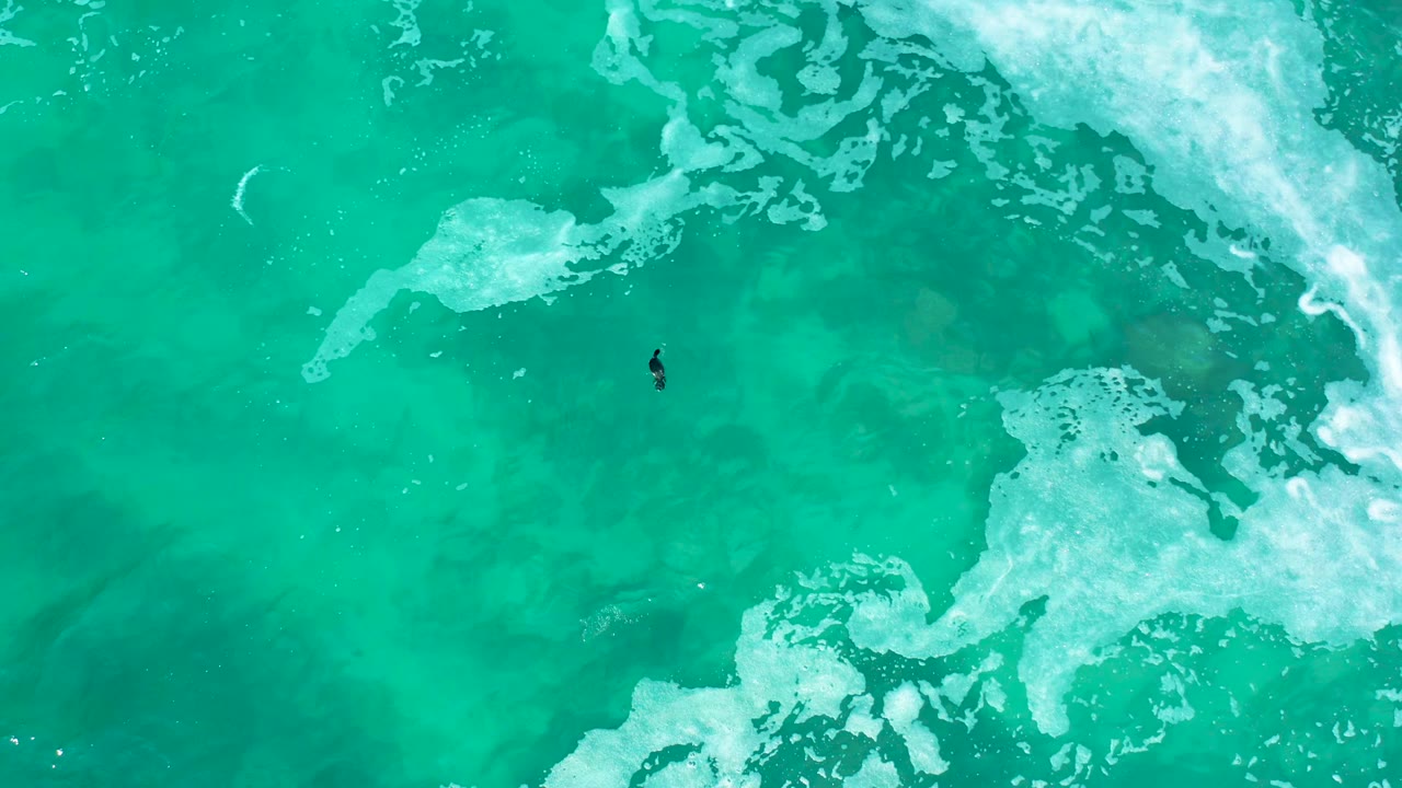 At Marquis Los Cabos, Mexico, an overhead view shows one small person in clear turquoise ocean water with white foam crossing the upper right and lower right edges.