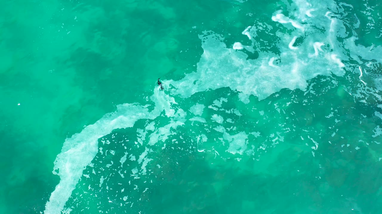 At Marquis Los Cabos, Mexico, white foam spreads diagonally through turquoise water while the small bodyboarder appears near the foamy break.