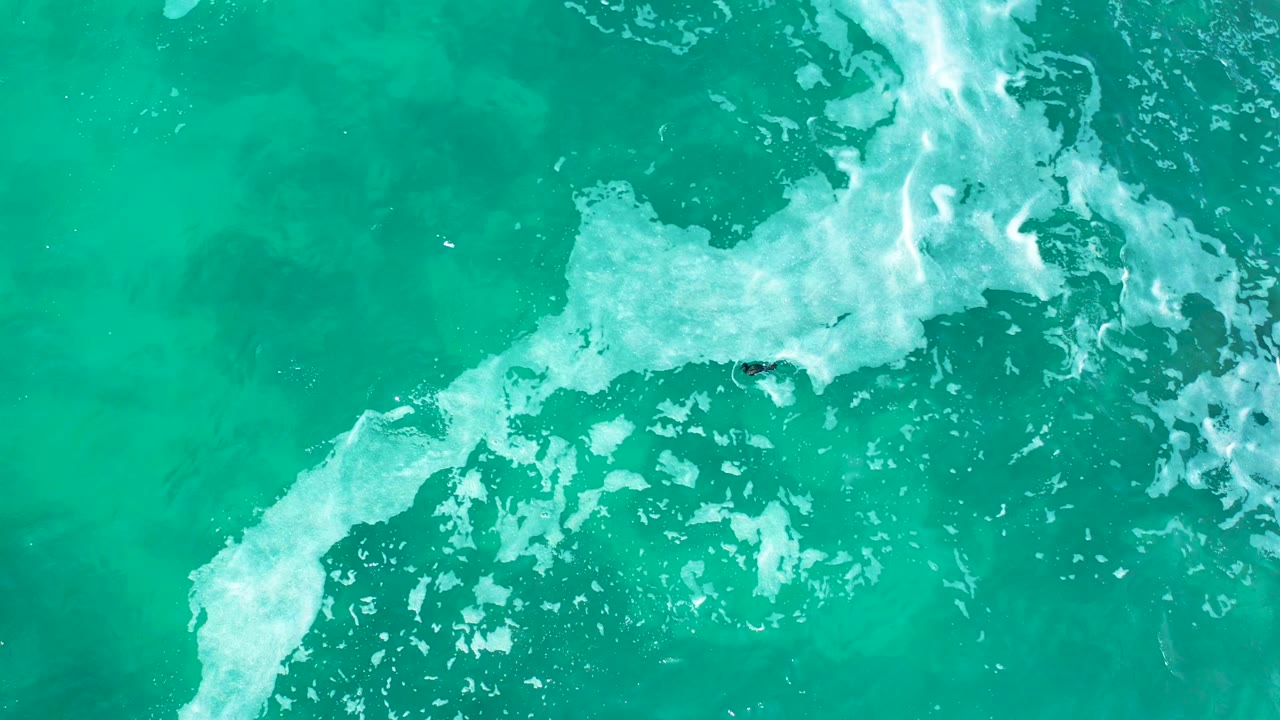 Overhead view at Marquis Los Cabos, Mexico showing turquoise water, a diagonal band of white foam, and a small distant surfer shape near the breaking wave with no recognizable people.
